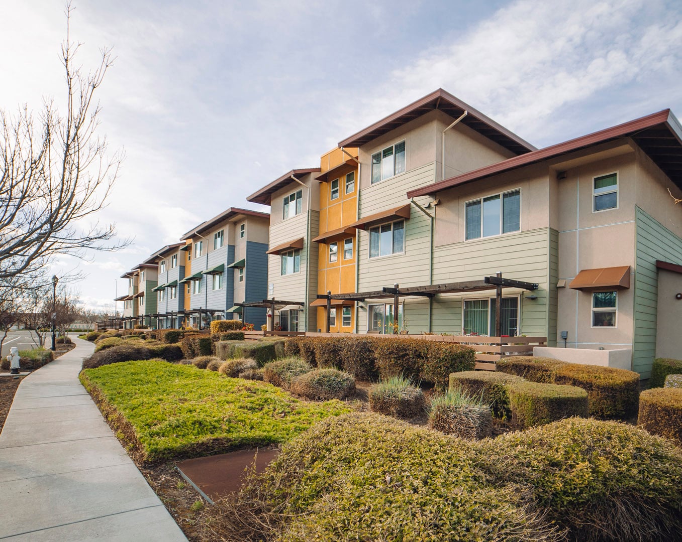 Modern three-story apartment building with beige and green siding, surrounded by neatly landscaped bushes and a sidewalk path, under a partly cloudy sky.